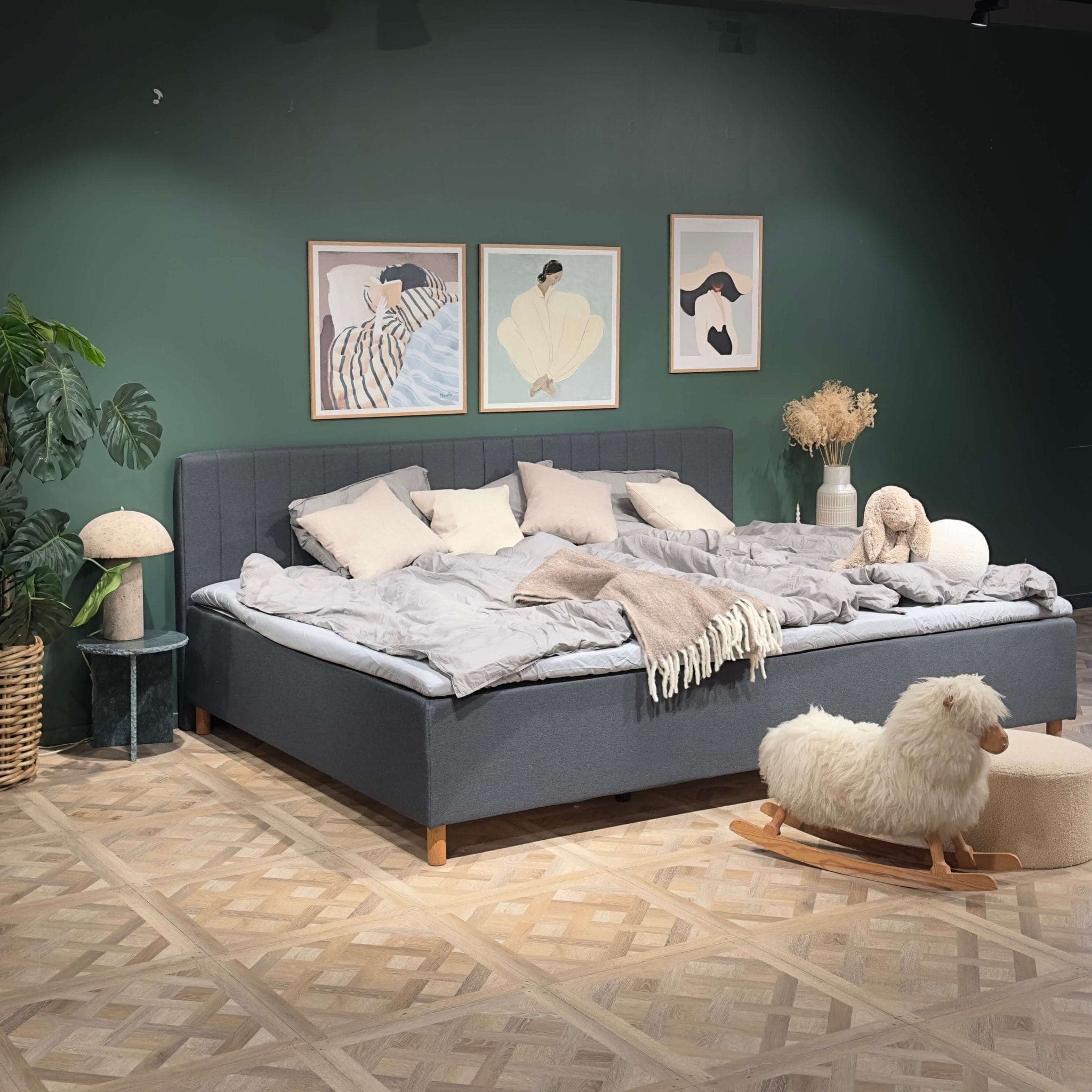 Breed, opgemaakt grijs familiebed in een slaapkamer met een groene muur en een schommelschaap op de vloer